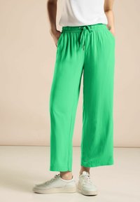 Zielone, lekkie spodnie z szerokimi nogawkami, posiadające elastyczny pas i sznurek do regulacji. Zawierają boczne kieszenie i gładką teksturę. Połączone z białymi trampkami.