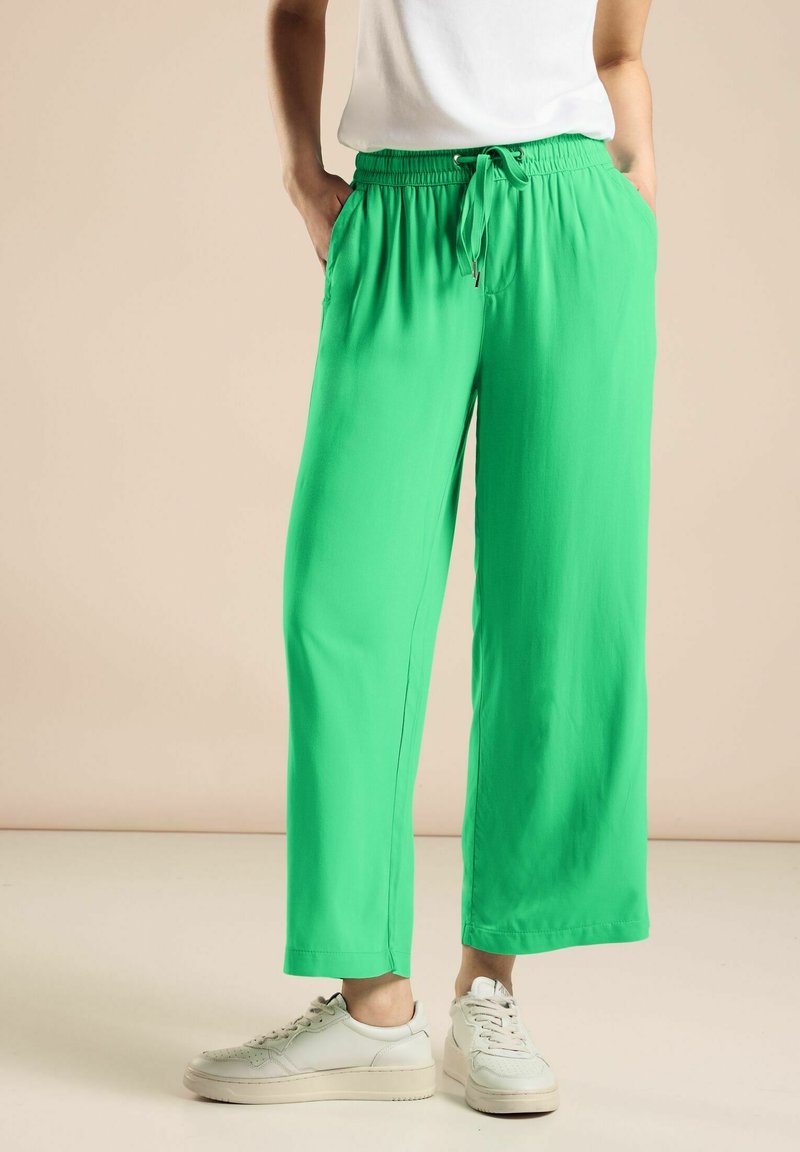 Zielone, lekkie spodnie z szerokimi nogawkami, posiadające elastyczny pas i sznurek do regulacji. Zawierają boczne kieszenie i gładką teksturę. Połączone z białymi trampkami.