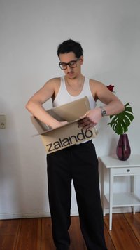 Moški v beli majici brez rokavov in črnih hlačah odpira škatlo Zalando v notranjosti, poleg bež mize s svetlo vijoličasto vazo in zeleno rastlino.