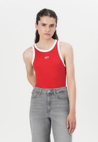 Rotes geripptes Tanktop mit weißem Rand, featuring ein zentrales "GAP"-Logo. Kombiniert mit hoch geschnittenen grauen Jeans.