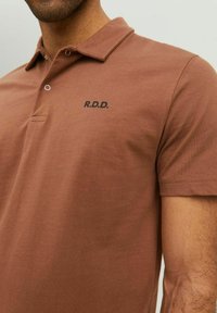 Hnedé polo tričko s golierom, krátkymi rukávmi a malým logom "R.D.D." v čiernej farbe. Má hladkú bavlnenú látku a dva gombíky pri krku.