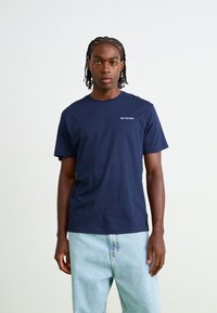 Marineblå bomulds T-shirt med korte ærmer og rund halsudskæring, med et lille hvidt Columbia-logo på venstre bryst. Lyse denimbukser.