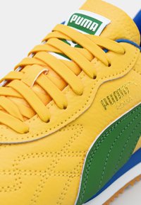 Αθλητικά παπούτσια PUMA με κίτρινο δέρμα στο επάνω μέρος, πράσινες λεπτομέρειες και επίπεδες κίτρινες κορδόνες. Ανυψωμένο λογότυπο και λεπτομέρεια υφής στην επιφάνεια.