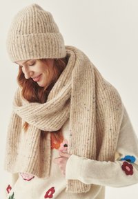 Bežová pletená čiapka a šál s vertikálnym žebrovaním, vyrobené z mäkké vlny, s farebnými škvrnami. Svetre majú kvetinové vyšívané akcenty.