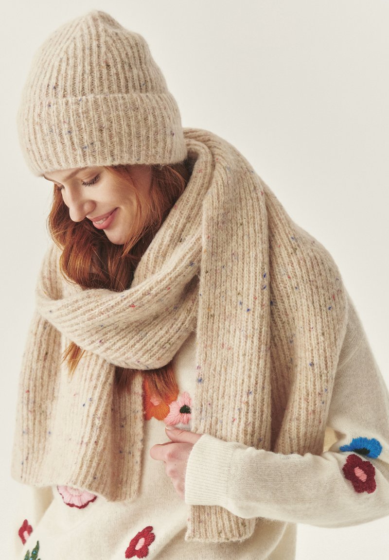 Bežová pletená čiapka a šál s vertikálnym žebrovaním, vyrobené z mäkké vlny, s farebnými škvrnami. Svetre majú kvetinové vyšívané akcenty.