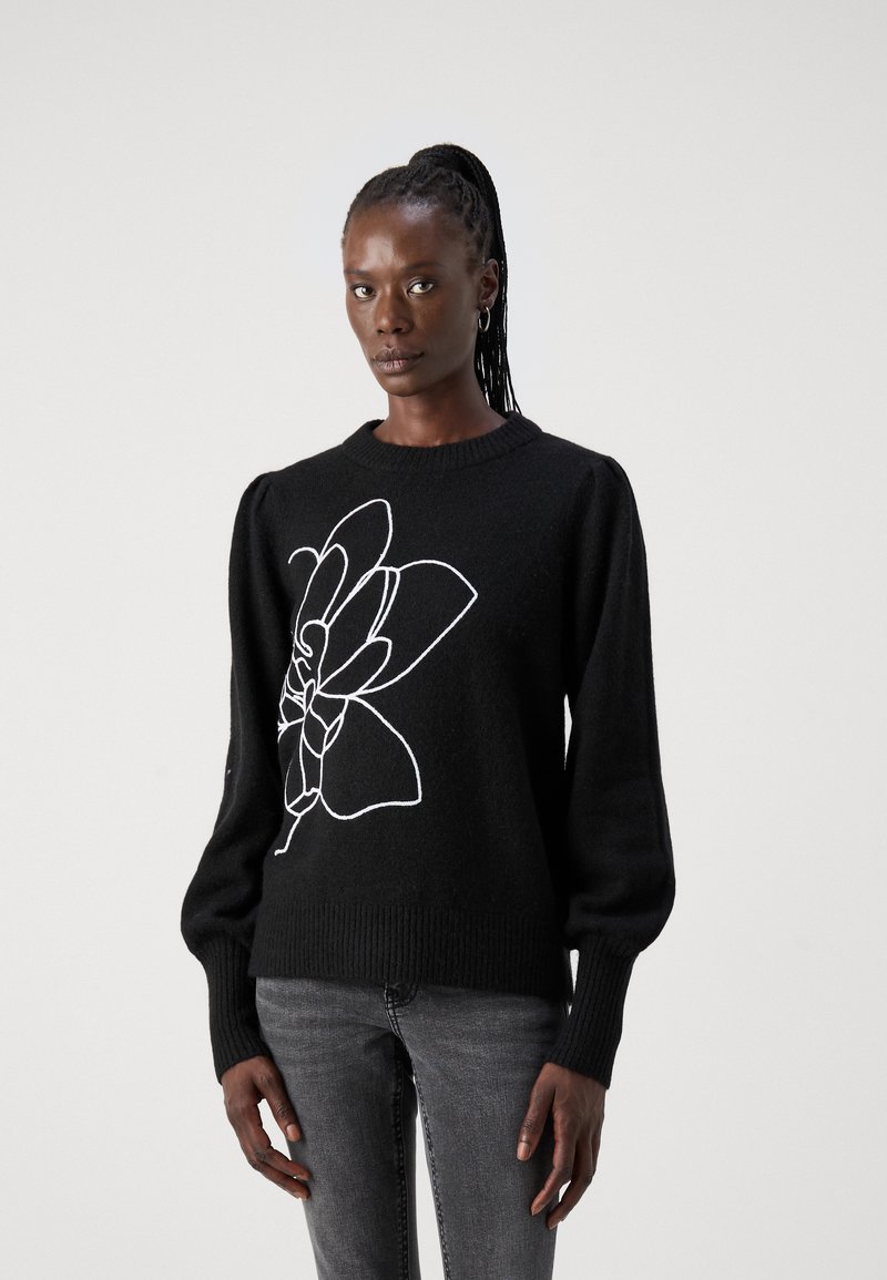 Pull en tricot noir avec de longues manches bouffantes et un ourlet côtelé, présentant un design floral blanc sur le devant. Porté avec un jean gris.