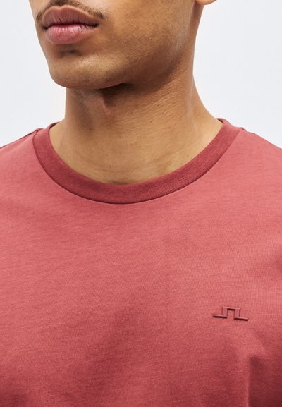 Nahaufnahme einer Person, die ein ausgewaschenes rosa T-Shirt mit Rundhalsausschnitt und einem kleinen, abstrakten Logo auf der linken Brust trägt.