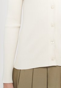 Cardigan blanc côtelé à manches longues avec boutons, associé à un pantalon plissé olive taille haute visible de la taille au poignet.