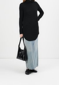 Žena v černém roláku a dlouhé světle modré sukni drží černou koženou kabelku s přezkou.