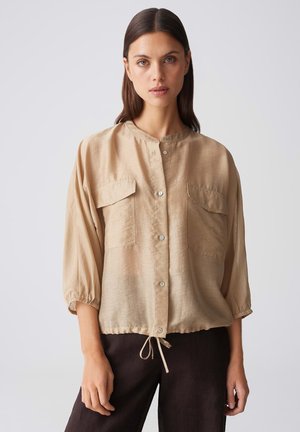 Vrouw met steil bruin haar die een beige blouse met knopen en een koordzoom draagt en zwarte broek, staande tegen een effen achtergrond.