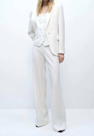 Femme portant un costume blanc avec un gilet croisé, un blazer et un pantalon à jambes larges, debout devant un fond clair uni.