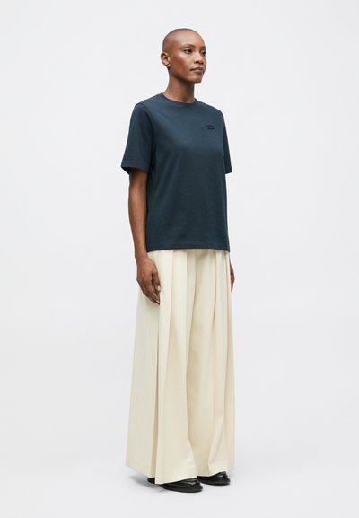 Námořnická modrá bavlněná tričko s krátkými rukávy má malou nášivku na hrudi. Kombinováno s krémovými, plisovanými kalhotami širokého střihu a černými botami.