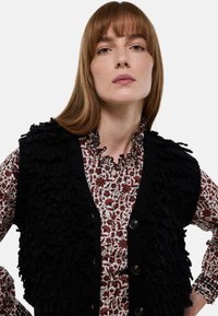Gilet noir avec des détails texturés et duveteux, porté par-dessus une chemise blanche avec un motif floral rouge. Comprend des boutons et un col à volants.