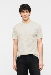 Νέος άνδρας με σγουρά μαλλιά, φορώντας μπεζ μπλουζάκι με μοτίβο και κείμενο, καθώς και μαύρο τζιν, όρθιος μπροστά σε απλό φόντο.