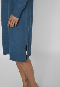 En kvinnas hand och ben syns, iklädd en blå stickad klänning med sidssprund, som visar en liten ankel-tatuering mot en ljusgrå bakgrund.