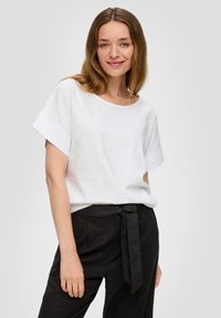 T-shirt en lin blanc avec un col rond large et des manches courtes, associé à un pantalon noir taille haute avec un détail de taille nouée.