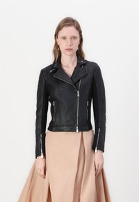 Giacca da moto in pelle nera con chiusura a zip asimmetrica, hardware argentato e finitura testurizzata. Abbinata a una gonna beige chiaro.