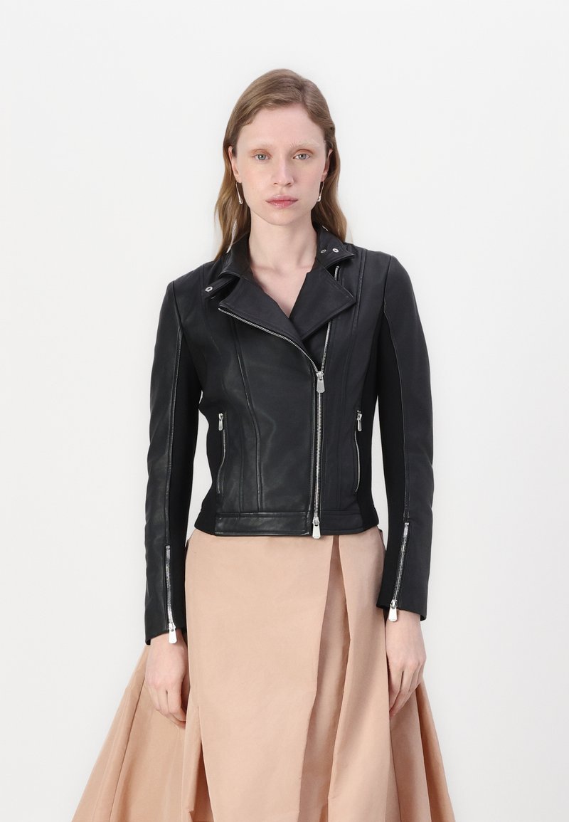 Giacca da moto in pelle nera con chiusura a zip asimmetrica, hardware argentato e finitura testurizzata. Abbinata a una gonna beige chiaro.