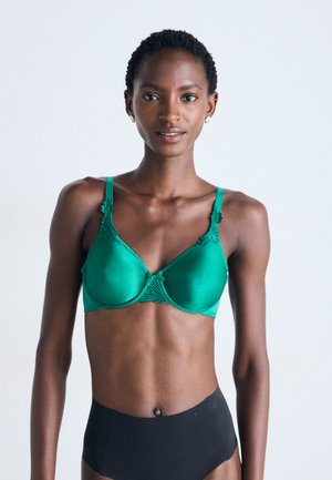 Slim woman with short curly hair wearing a green bra with lace details and high-waisted black underwear against a plain background.