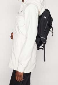 Giacca impermeabile bianca con cappuccio, dotata di vita elastica, e zaino nero con spallacci regolabili. Etichetta "DryVent" visibile sulla manica.