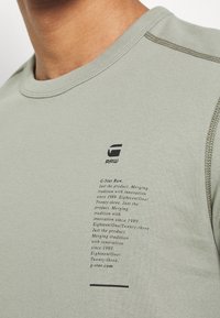 Hellgrünes Baumwoll-T-Shirt mit Rundhalsausschnitt. Auf der linken Seite befinden sich schwarze gedruckte Markenlogos und Texte. Glatte Textur mit kontrastierenden Nähten.