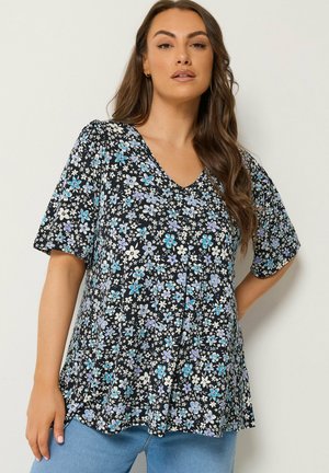 Femme portant un chemisier noir à col en V avec un motif floral bleu, blanc et violet et des manches courtes, associé à un jean bleu clair.