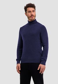 Granatowy sweter z golfem wykonany z teksturowanej dzianiny, z długimi rękawami oraz ściągaczami na dole i mankietach, noszony w połączeniu z ciemnymi spodniami.