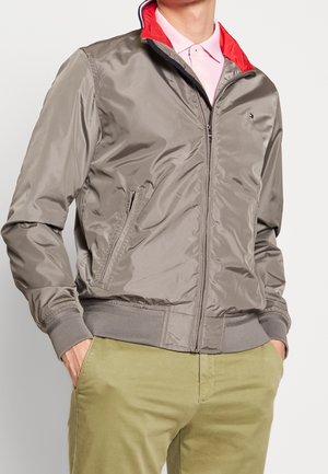 Homme portant une veste taupe zippée avec doublure rouge au col, chemise rose clair et pantalon vert clair, les mains dans les poches.