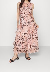 Maximekkoin pastellin vaaleanpunaisena, jossa on höyhenmäinen kuvio, varustettuna röyhelöhihoilla, soljuvalla siluetilla ja mustilla remmisandaaleilla.