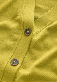 Sweter w kolorze jasnopomarańczowym, z dekoltem w kształcie litery V i dwoma okrągłymi, marmurkowymi guzikami z przodu. Miękka faktura z prążkowanymi detalami.