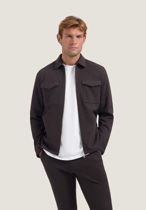 Man draagt een donkerbruine jas met rits en klepzakken over een wit T-shirt en bijpassende donkere broek, staand met handen in de zakken.