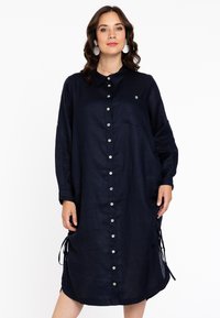 Marineblaues Leinenkleid mit Kragen, durchgehender Knopfleiste, langen Ärmeln und seitlichen Bindebändern. Mit einer Fronttasche und glänzenden Knopfakzenten.