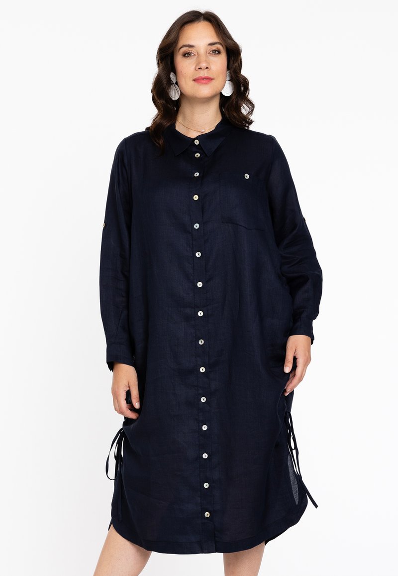 Marineblaues Leinenkleid mit Kragen, durchgehender Knopfleiste, langen Ärmeln und seitlichen Bindebändern. Mit einer Fronttasche und glänzenden Knopfakzenten.