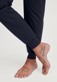 Pantalons de jogging marine avec des poignets élastiques, associés à un pied nu mettant en avant des tatouages, soulignant le ton et la texture de la peau.