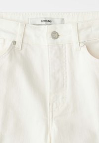 Nahaufnahme des Taillenbereichs einer weißen Jeans aus Denim mit silbernem Knopf und Gürtelschlaufen, versehen mit einem Etikett mit der Aufschrift „someday“.