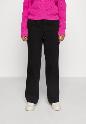 Personne portant un pull en tricot rose vif, un pantalon noir ample et des baskets blanches, debout devant un fond blanc uni.