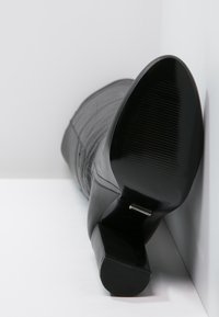Schwarze High-Heel-Stiefelette mit einem strukturierten Lederoberteil, runder Zehenform und einer gerippten Gummisohle. Der Absatz ist dick und konisch geformt für Stabilität.