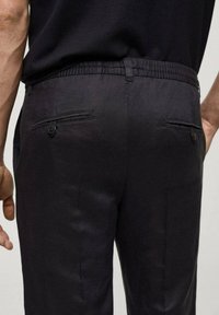 Black trousers with an elastic waistband, featuring two back pockets with button closures and a smooth, lightweight texture.