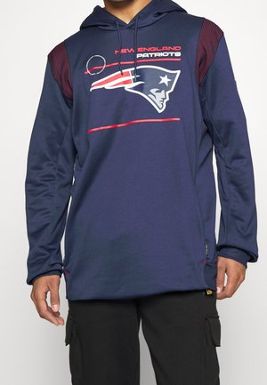 Sweat à capuche bleu marine avec accents rouges, arborant le logo et le texte des New England Patriots, porté avec un pantalon cargo noir.