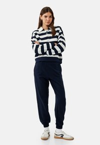 Gestreifter Pullover in Marineblau und Weiß mit Kragen und Knopf. Kombiniert mit Marineblauem Jogginghose und weißen Turnschuhen mit braunen Akzenten.