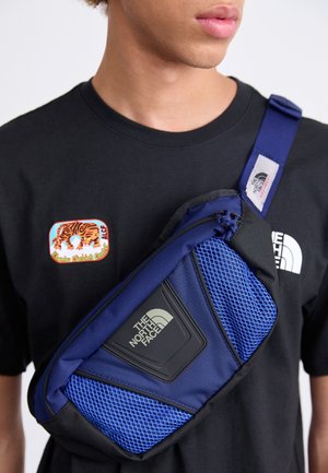 Jong persoon draagt een zwart T-shirt met een tijgerpatch en een blauwe The North Face crossbody tas over de borst.