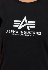 Čierne bavlnené tričko s bielym logom "Alpha Industries" s ostrým dizajnom a horizontálnymi pásikmi dole, so textom indikujúcim umiestnenie.
