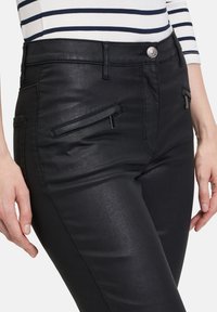 Schwarze Fake-Lederhosen mit glatter Textur, ausgestattet mit Fronttaschen und metallischen Akzenten an den Reißverschlüssen und dem Knopf.