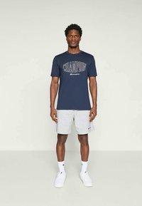 Marinblå bomull t-shirt med textdesign "CHAMPION", kombinerad med ljusgrå shorts, vita strumpor och vita sneakers; modellen står mot en enfärgad bakgrund.