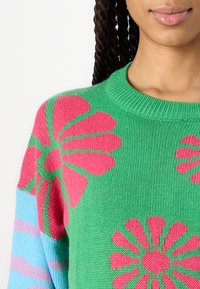 Pull en tricot avec une base verte, de grands motifs floraux roses et des manches rayées bleues ; col rond côtelé et matière texturée.