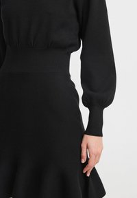Svart klänning med en figursydd överdel, puffärmar, ett ribbat midjeband och en utsvängd fåll som betonas av ett strukturerat stickat tyg.