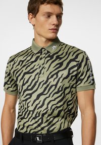 Grön pikétröja med ett svart tigerstrimmönster, med krage, tre knappar och en logotyp på ärmen. Tyget verkar slätt.