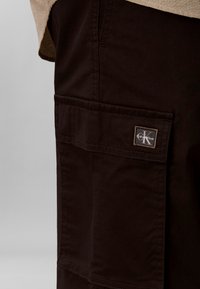 Gros plan d'une poche de pantalon marron foncé avec un écusson logo Calvin Klein cousu sur le rabat de la poche.