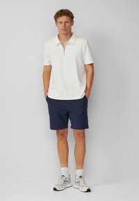 Hellbeige Polo-Shirt mit einem Reißverschlusskragen, kombiniert mit navy Shorts. Die Schuhe haben eine Mischung aus grauen und weißen Akzenten. Lässiges Design.
