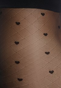 Collant trasparenti marroni con un motivo a diamante, decorati con piccoli cuori neri. Texture morbida e cintura nera rinforzata.